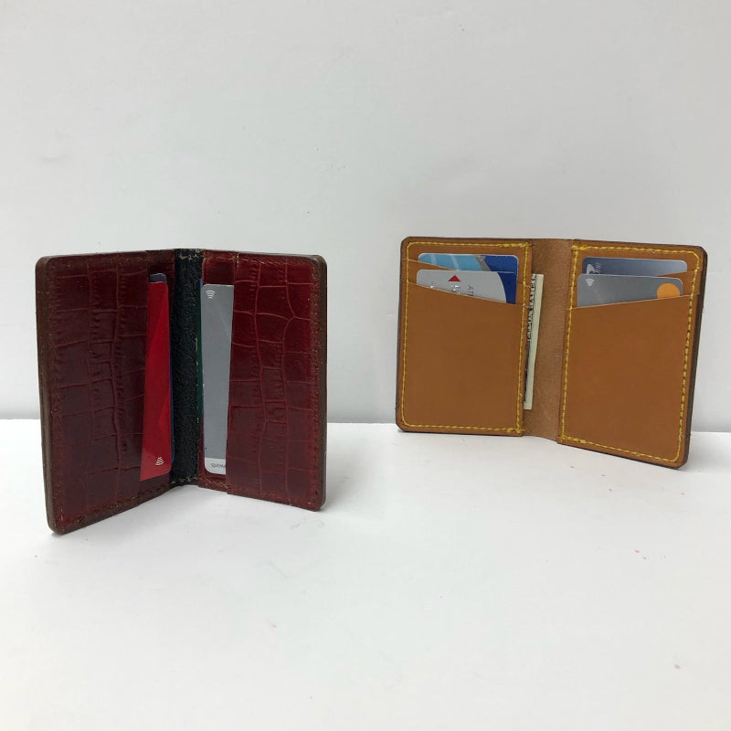 Two slim leather cardholder wallets shown open on a white surface against a white wall. The wallet on the left is dark burgundy with a crocodile-embossed texture and a red interior; the wallet on the right is tan with a smooth interior and visible card slots.