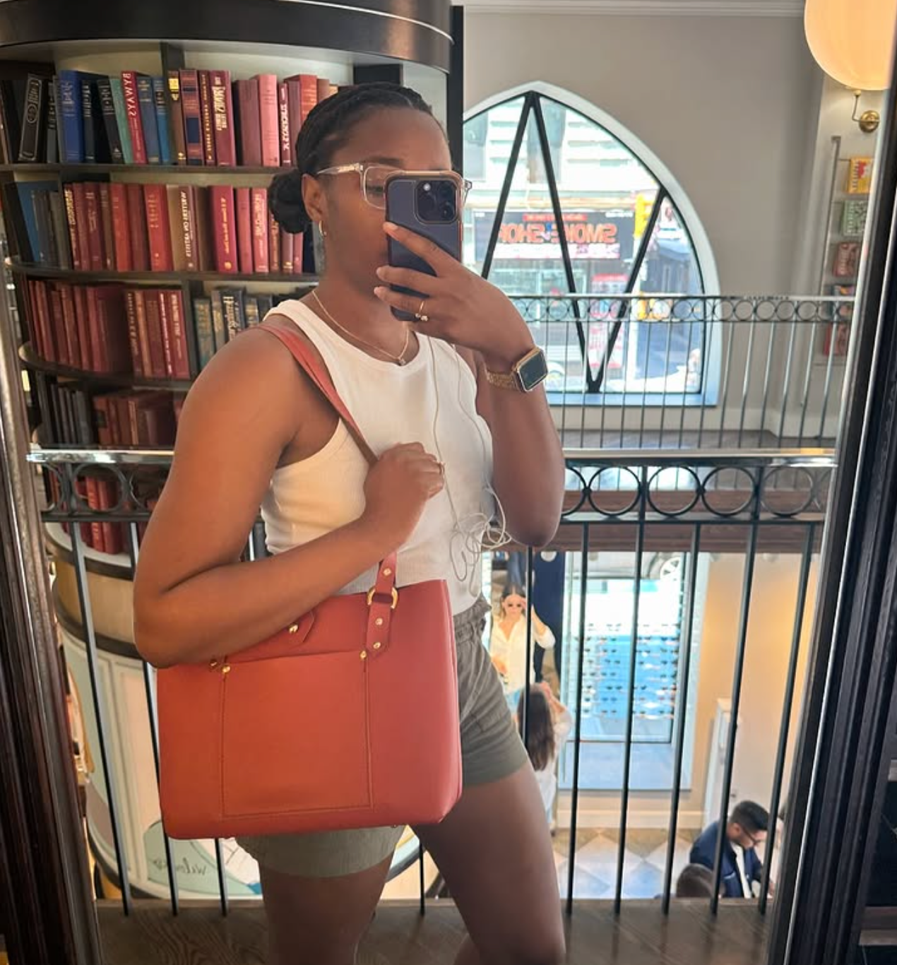 Person wearing a leather tote taking a mirror selfie in a bookstore with a bookshelf and cityscape in the background.